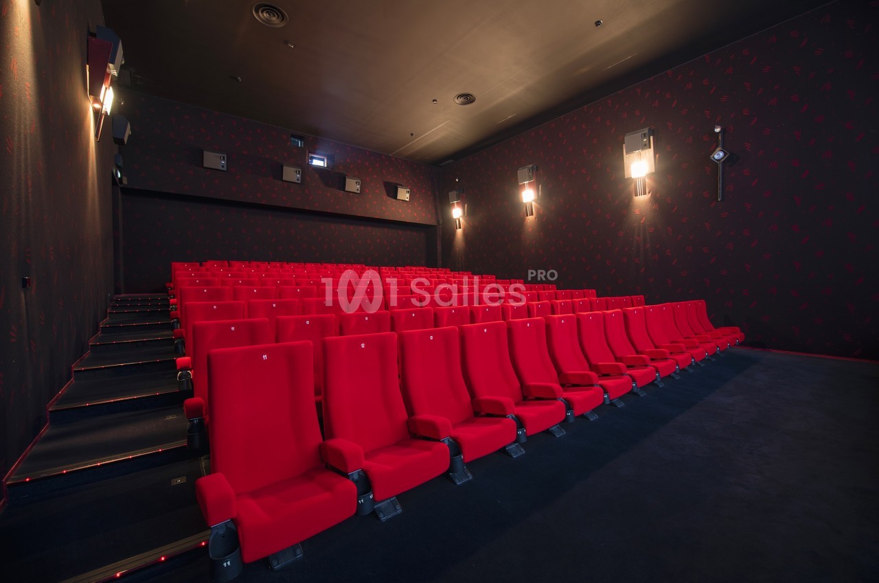 Salle de cinéma vide avec des rangées de fauteuils rouges et des murs sombres éclairés par des appliques murales.
