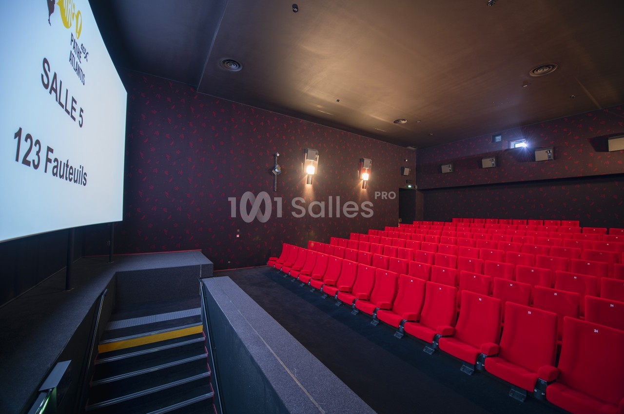 Salle de cinéma vide avec des rangées de fauteuils rouges, écran blanc et éclairage tamisé.