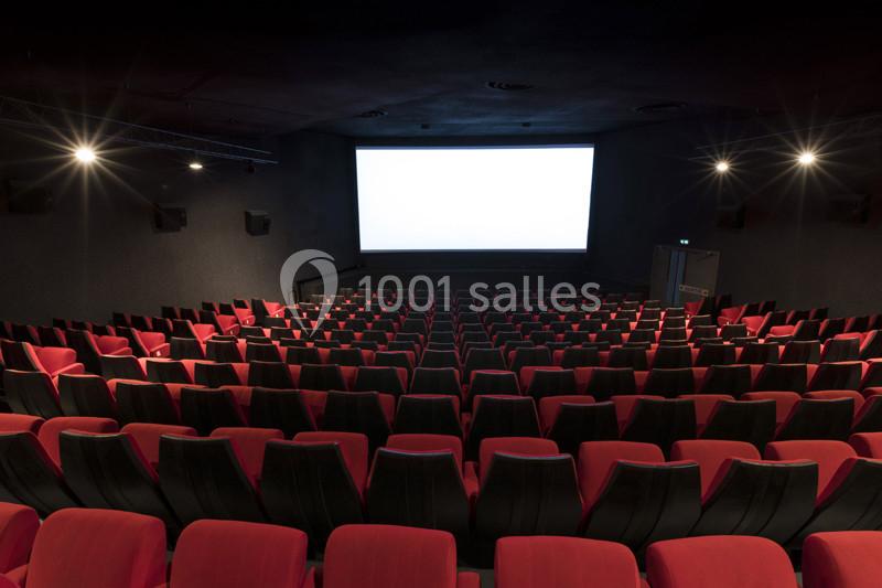 Salle de cinéma vide avec des rangées de fauteuils rouges et un grand écran blanc allumé.
