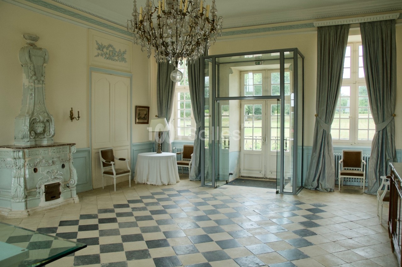 Salle lumineuse avec carrelage en damier, cheminée ancienne, lustre et grandes fenêtres donnant sur un jardin.