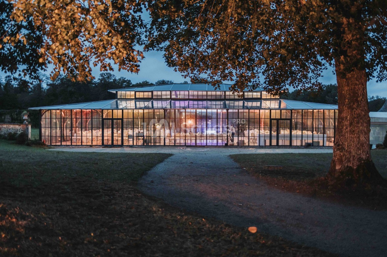 Grande verrière illuminée en soirée, entourée d'arbres et située dans un environnement naturel.