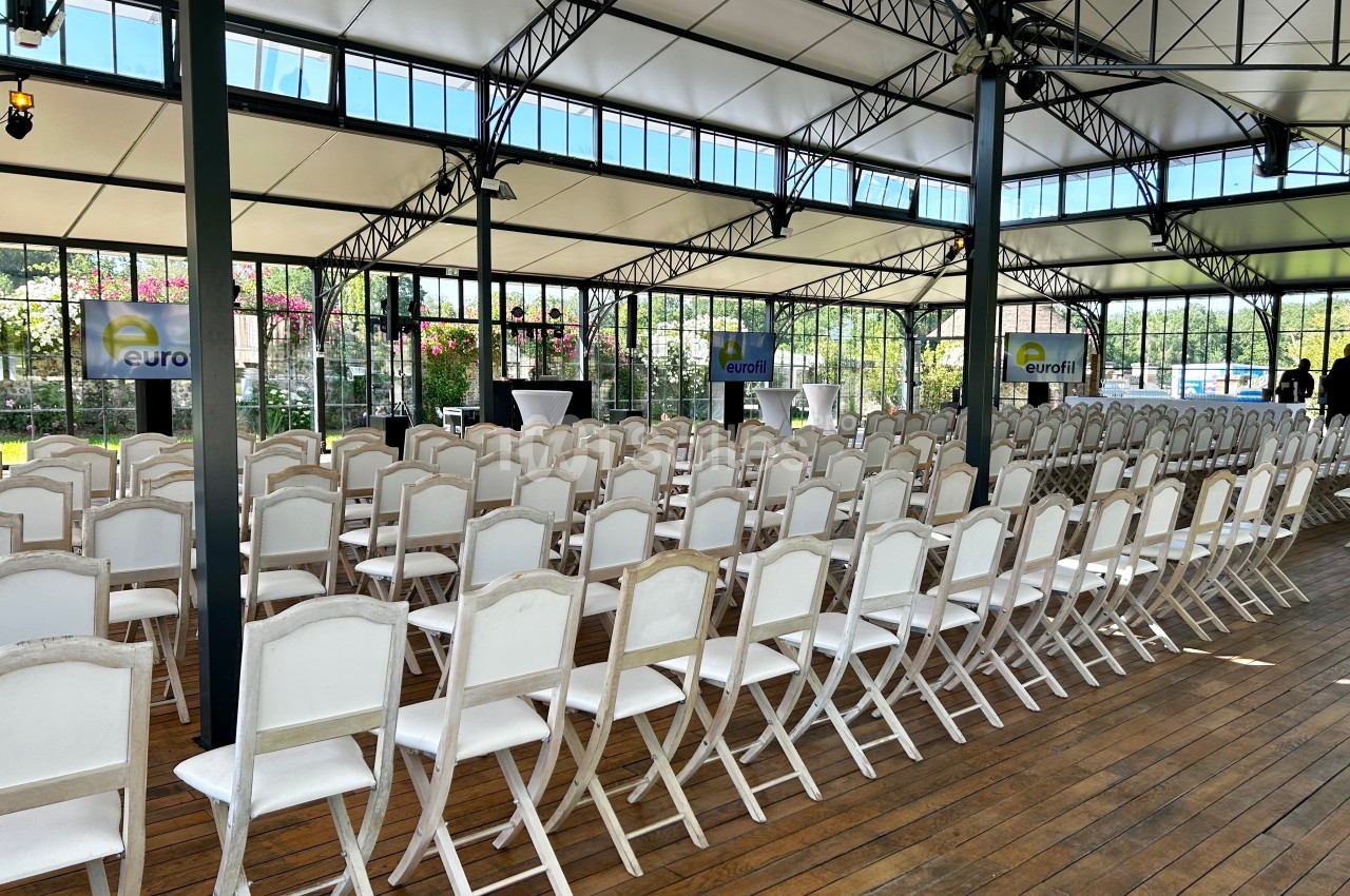 Salle lumineuse avec des rangées de chaises blanches alignées, prête pour un événement ou une conférence.