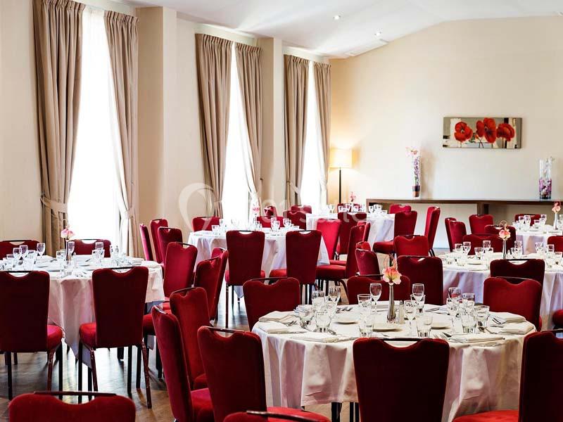 Salle de réception avec tables rondes dressées, nappes blanches, chaises rouges et lumière naturelle.