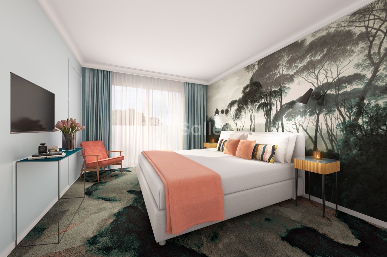 Chambre lumineuse avec un lit double, un mur décoré de motifs forestiers, un fauteuil et une grande fenêtre avec rideaux.