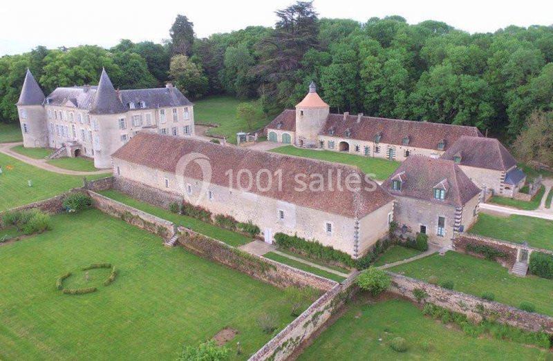 Vue aérienne d'un château entouré de bâtiments annexes, de jardins et de pelouses verdoyantes, bordé par une forêt.