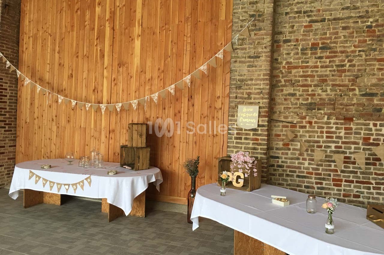 Salle décorée pour un événement avec des tables recouvertes de nappes blanches, guirlandes et éléments rustiques en bois.