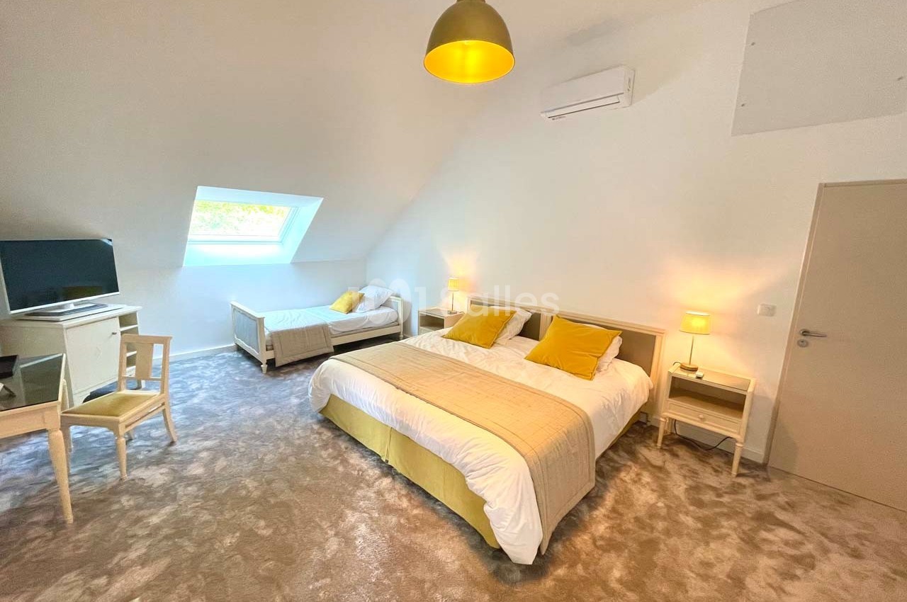Chambre lumineuse sous combles avec lit double, lit simple, bureau, télévision, lampes de chevet et moquette grise.