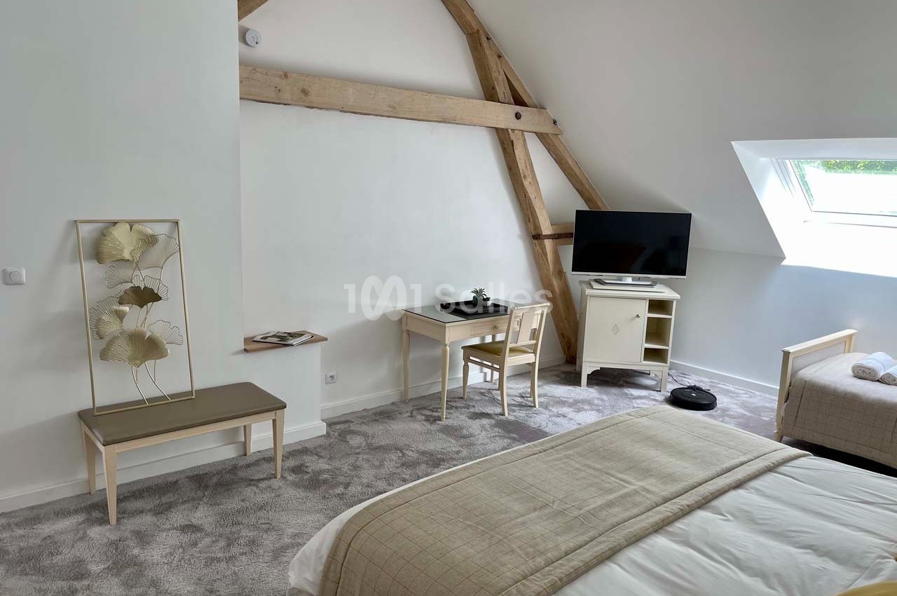 Chambre lumineuse sous combles avec poutres apparentes, lit double, bureau, télévision et fenêtre de toit.