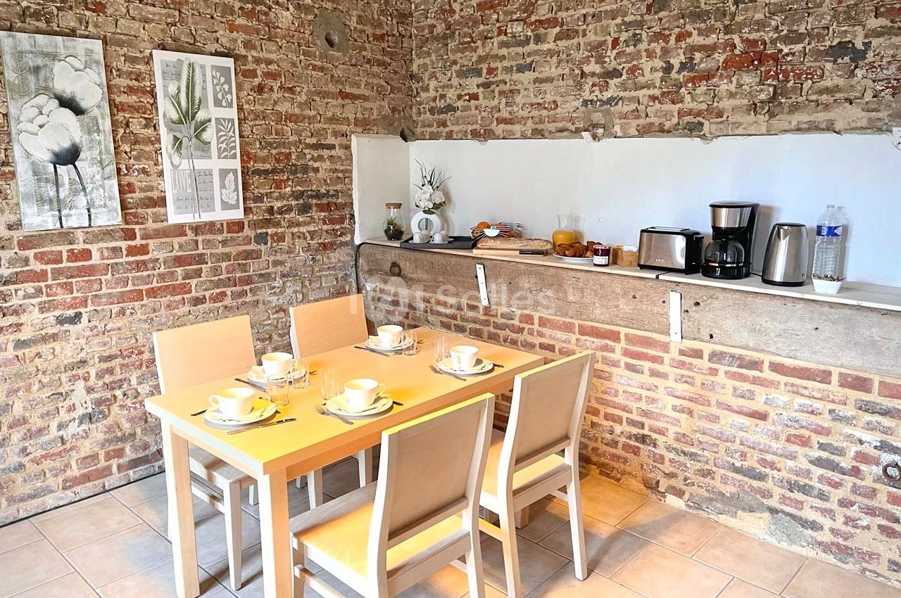 Salle à manger avec table en bois, chaises, vaisselle disposée et mur en briques apparentes.
