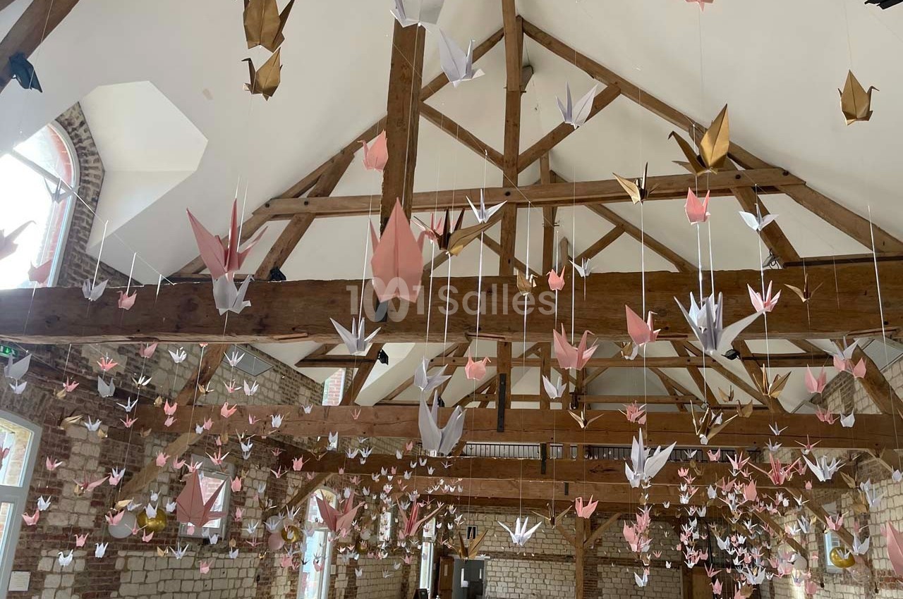 Origamis de grues suspendus au plafond d'une salle avec charpente en bois et murs en briques apparentes.