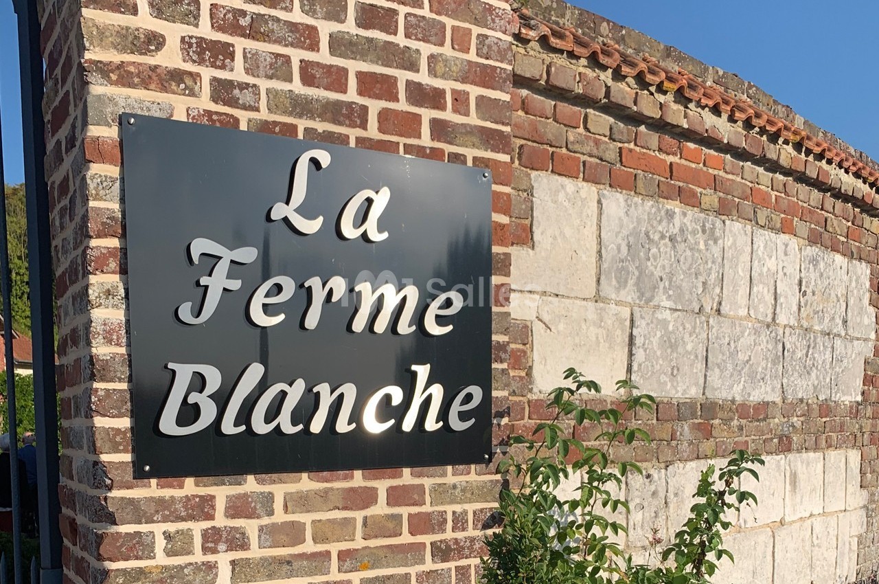 Panneau noir avec l'inscription ’La Ferme Blanche’ fixé sur un mur en briques et pierres sous un ciel bleu.