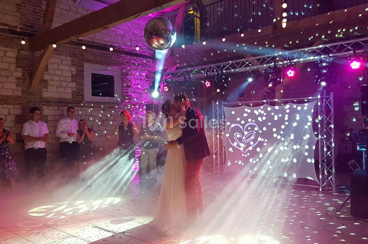 Un couple danse sous une boule à facettes dans une salle éclairée par des lumières colorées, entouré d'invités.