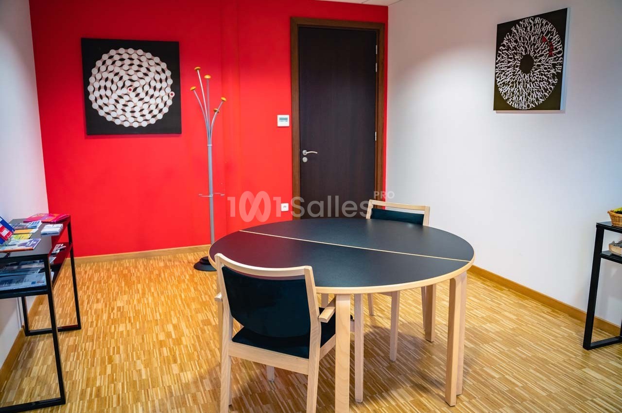 Salle avec deux chaises autour d'une table ronde, murs rouges et blancs décorés de tableaux géométriques.