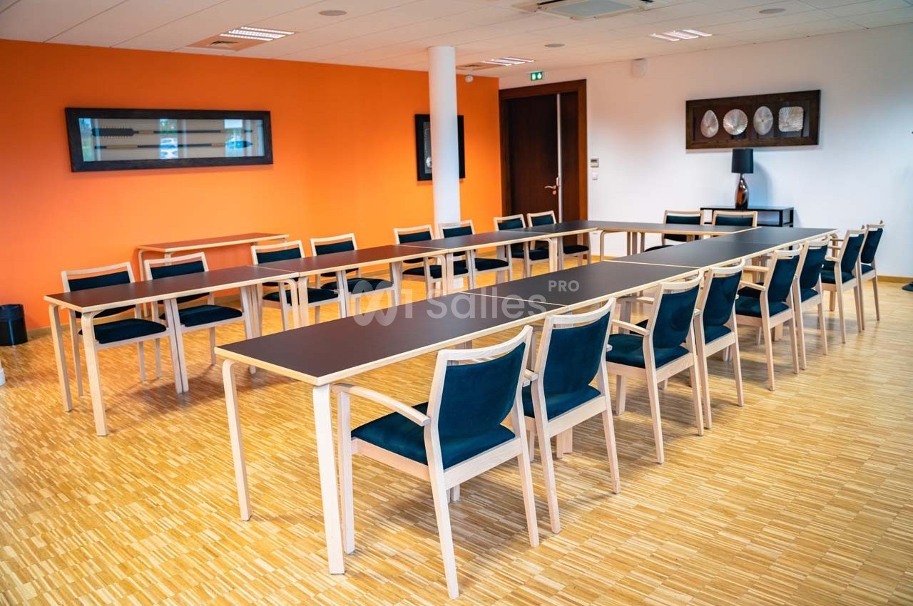 Salle de réunion lumineuse avec une grande table en U, des chaises en bois et un mur orange en arrière-plan.
