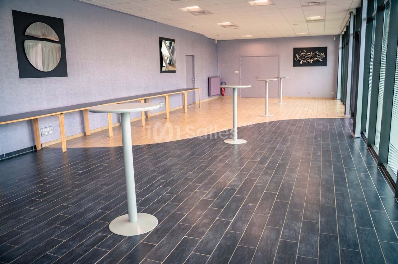 Salle vide avec sol en carrelage et parquet, tables hautes, murs violets et grandes fenêtres laissant entrer la lumière.
