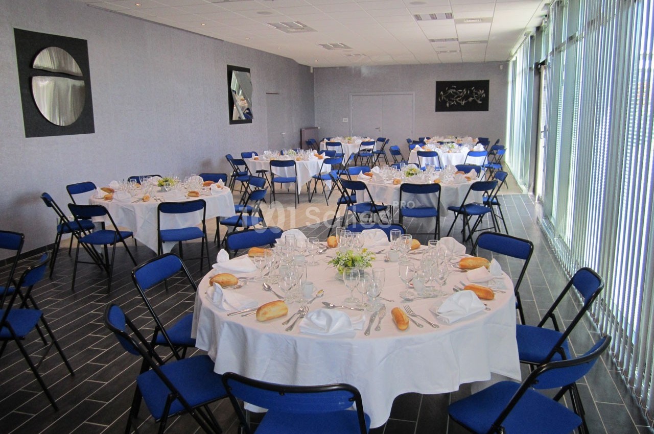 Salle de réception avec tables rondes dressées, nappes blanches, chaises bleues et lumière naturelle entrant par des baies…
