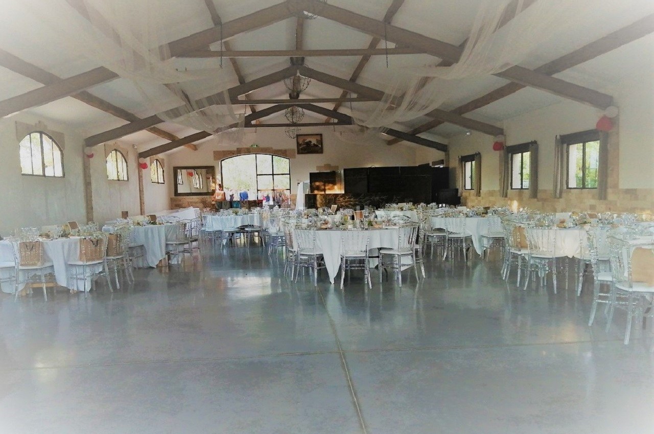 Salle de réception décorée avec des tables rondes et rectangulaires dressées, sous une charpente en bois apparente.