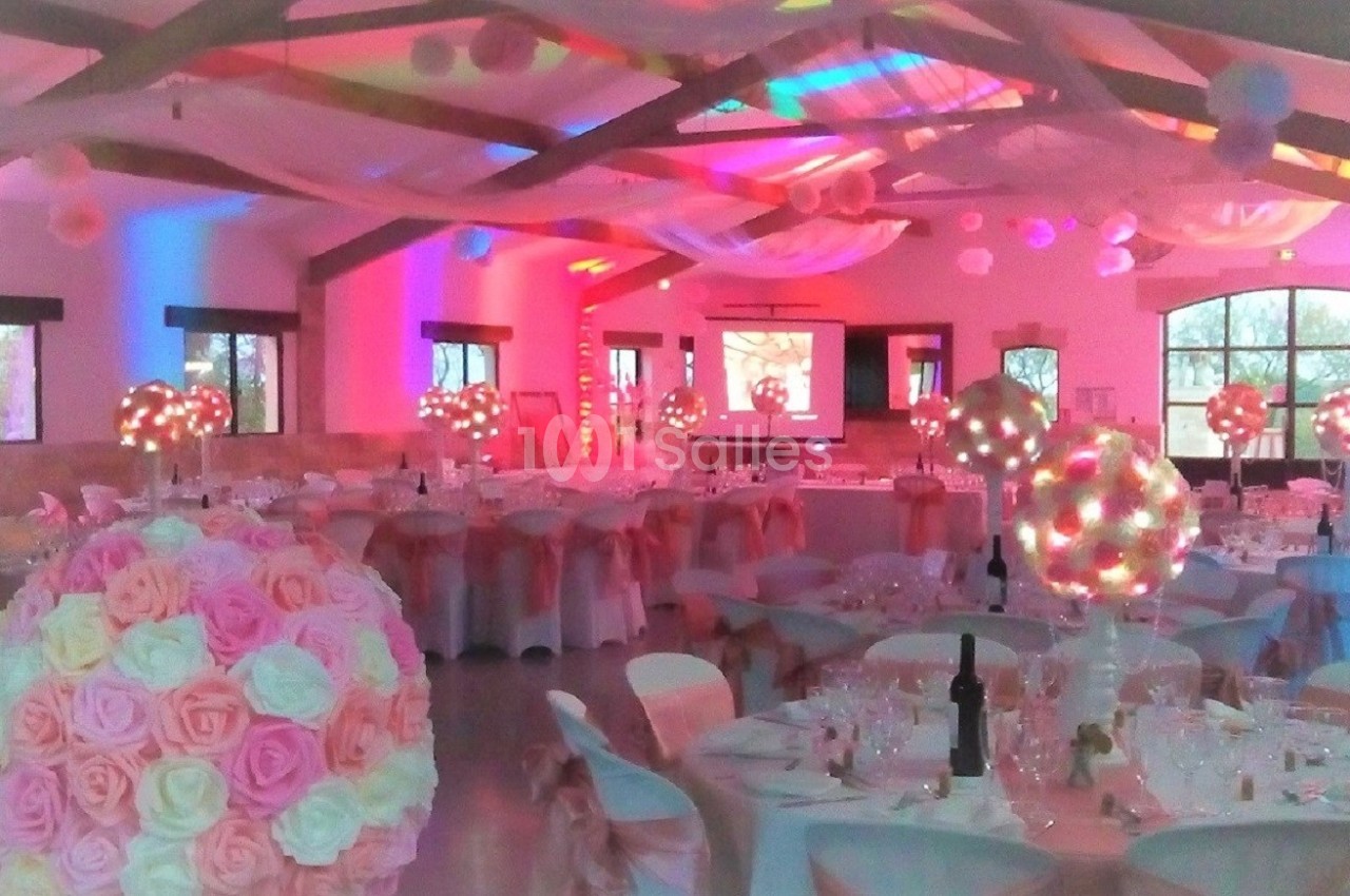 Salle de réception décorée avec des tables dressées, des guirlandes lumineuses et des arrangements floraux roses.