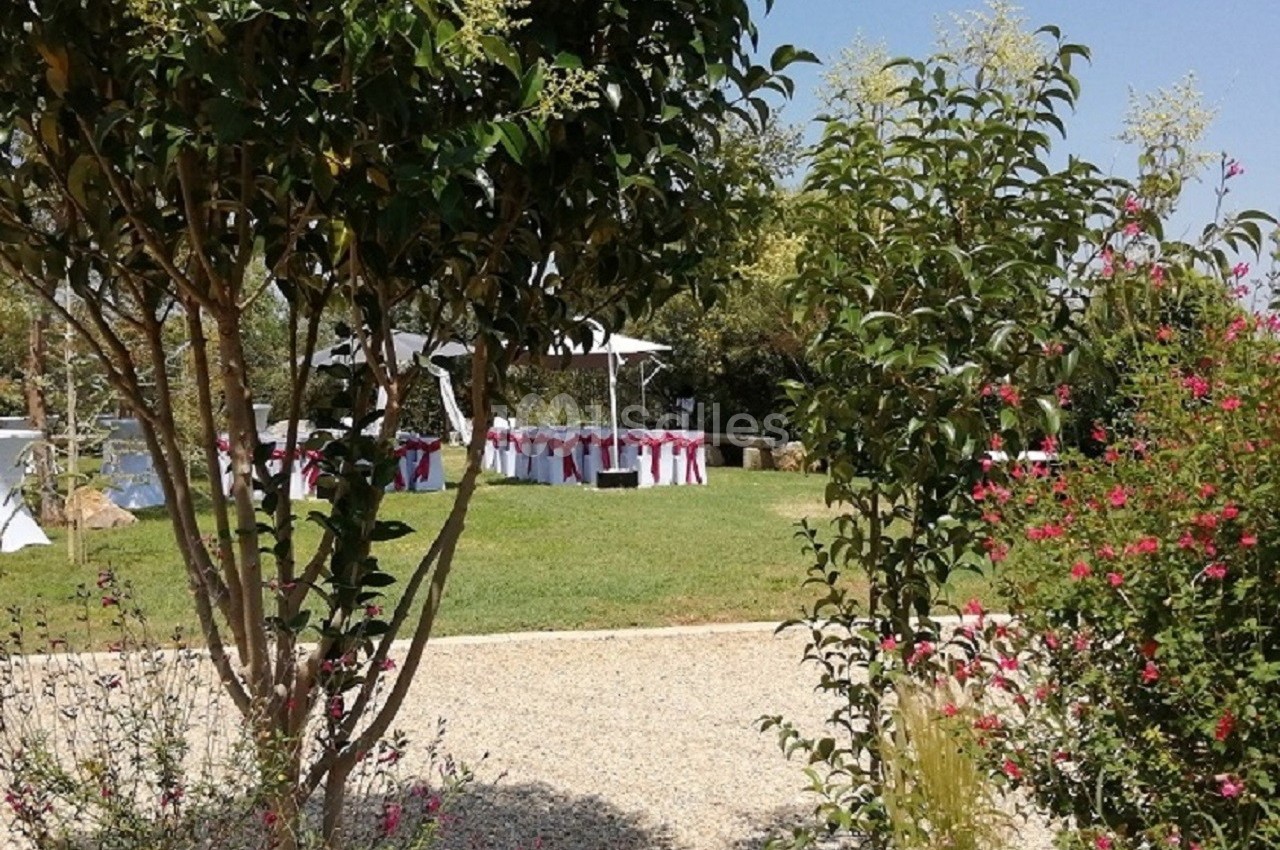 Jardin avec pelouse, arbres et tables décorées de nappes blanches et rubans rouges, sous un ciel dégagé.