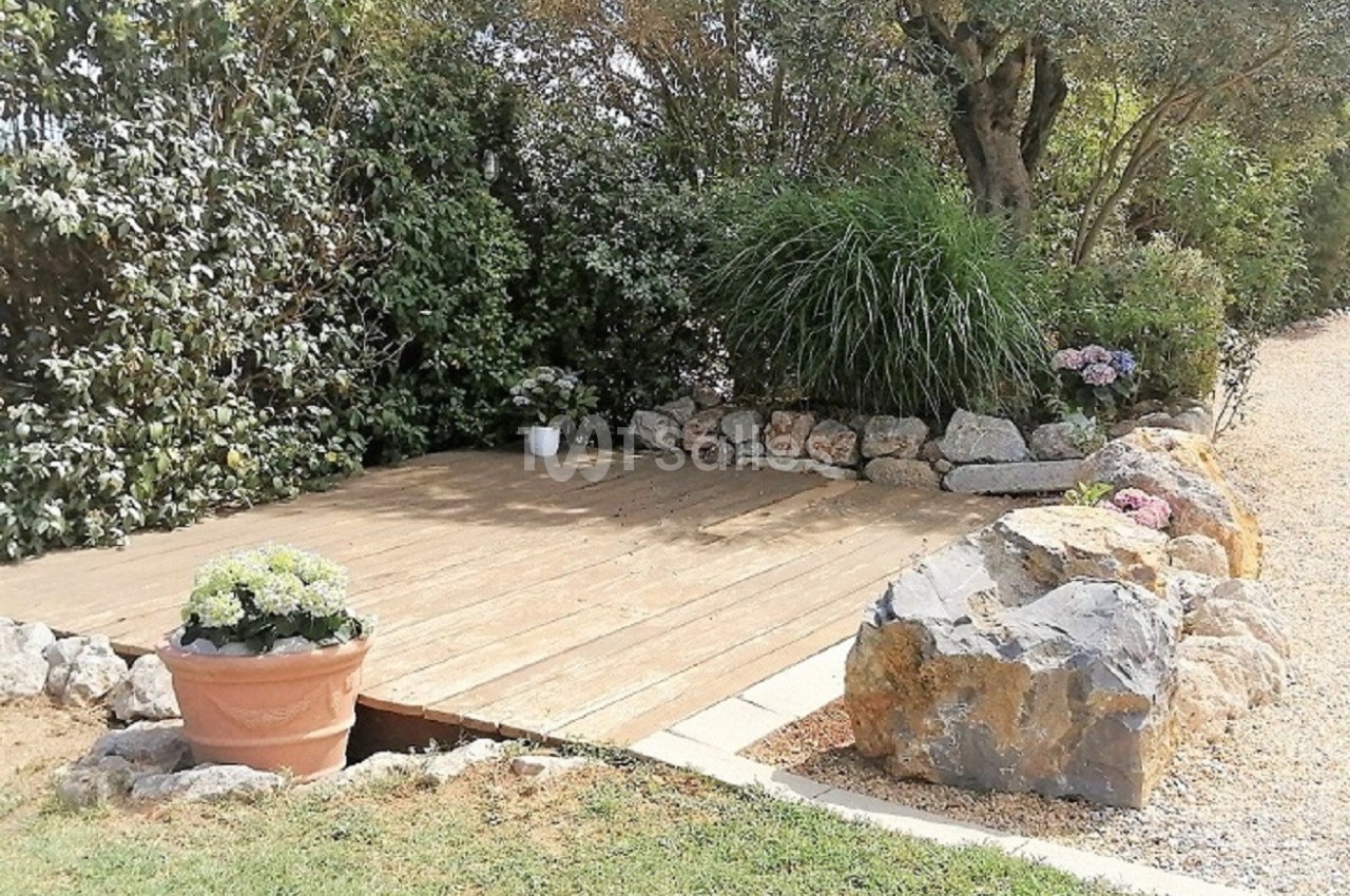 Terrasse en bois entourée de plantes, buissons et rochers, avec un pot de fleurs au premier plan.