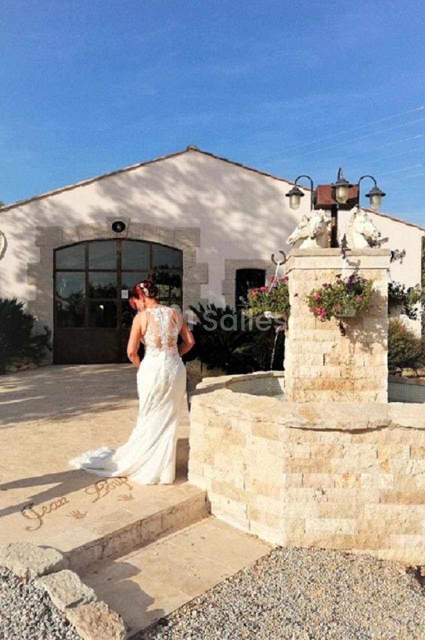 Une mariée en robe blanche marche près d'une fontaine en pierre devant un bâtiment lumineux sous un ciel bleu.