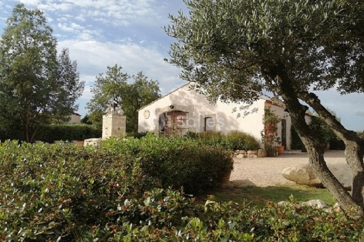 Maison blanche entourée de végétation méditerranéenne, avec un arbre au premier plan et un ciel partiellement nuageux.
