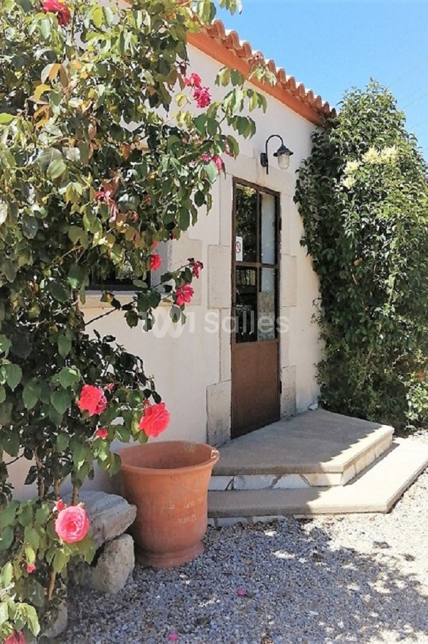 Façade d'une petite maison en pierre avec porte en bois, entourée de rosiers et d'un pot en terre cuite.