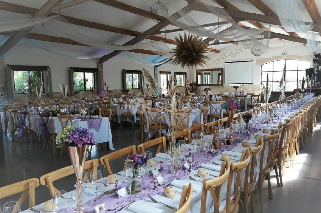 Salle de réception décorée pour un événement, avec tables alignées, nappes violettes, fleurs et chaises en bois.