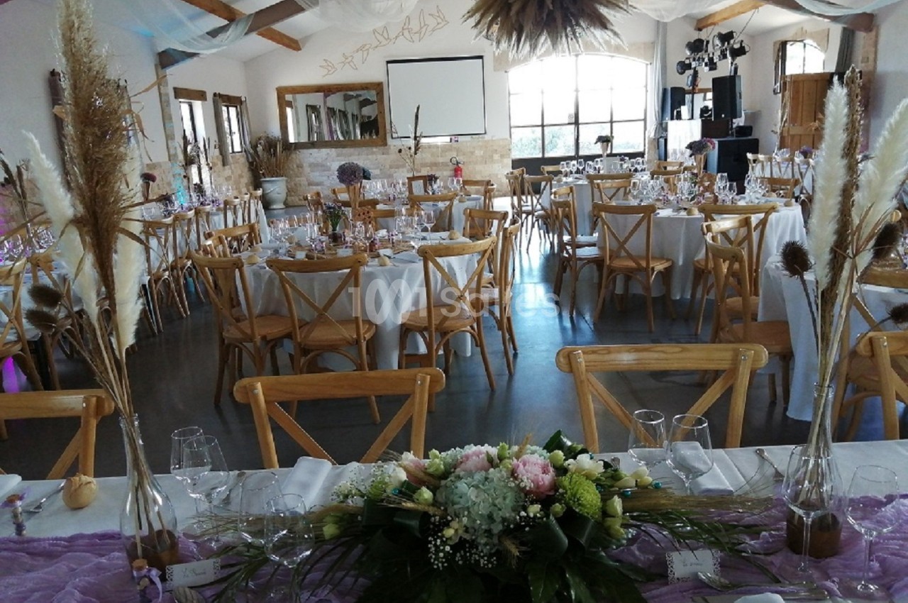 Salle de réception décorée pour un événement, avec tables dressées, chaises en bois et compositions florales.