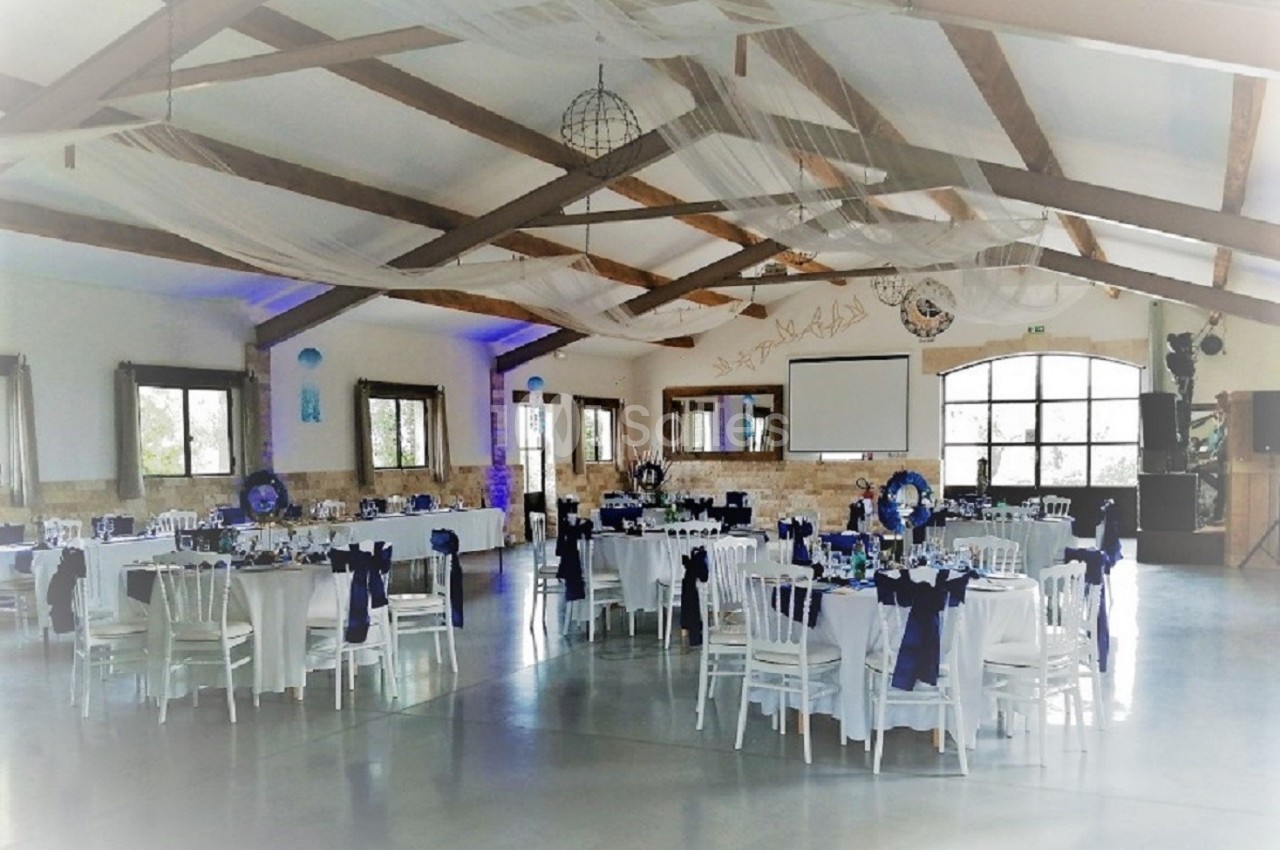 Salle de réception décorée avec des tables rondes dressées, nappes blanches et chaises ornées de rubans bleus.