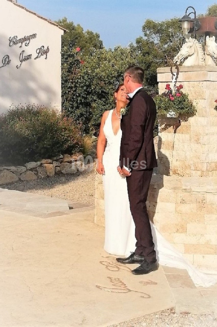 Un couple en tenue de mariage pose près d'une fontaine en pierre devant un bâtiment blanc.
