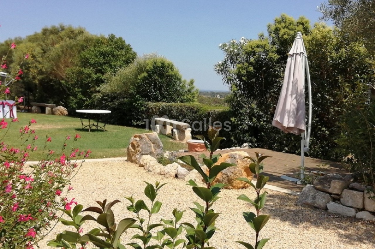 Jardin avec terrasse en gravier, parasol fermé, table en arrière-plan et végétation luxuriante sous un ciel dégagé.