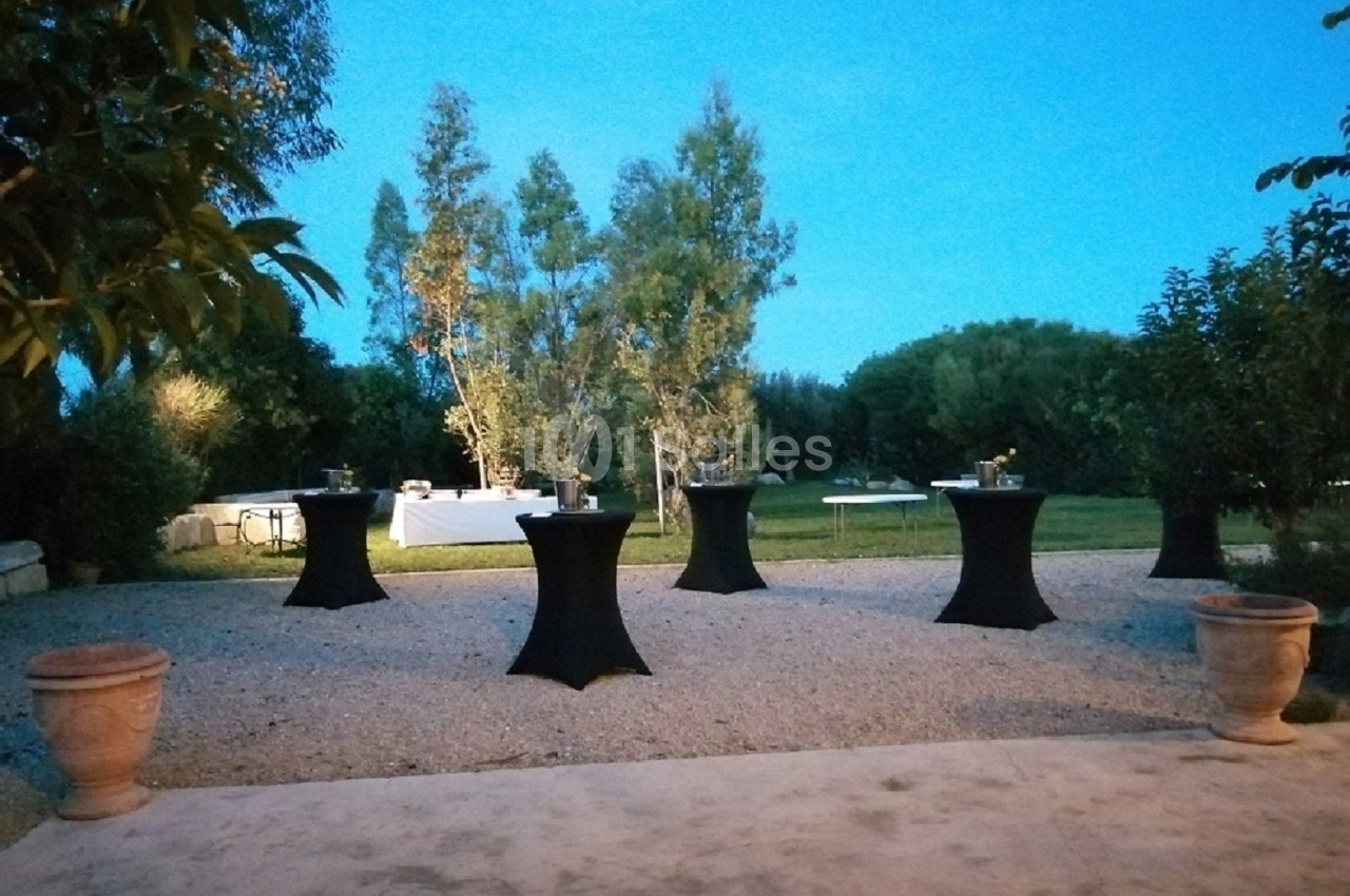 Espace extérieur aménagé avec des tables hautes noires sur une terrasse gravillonnée, entouré de verdure au crépuscule.
