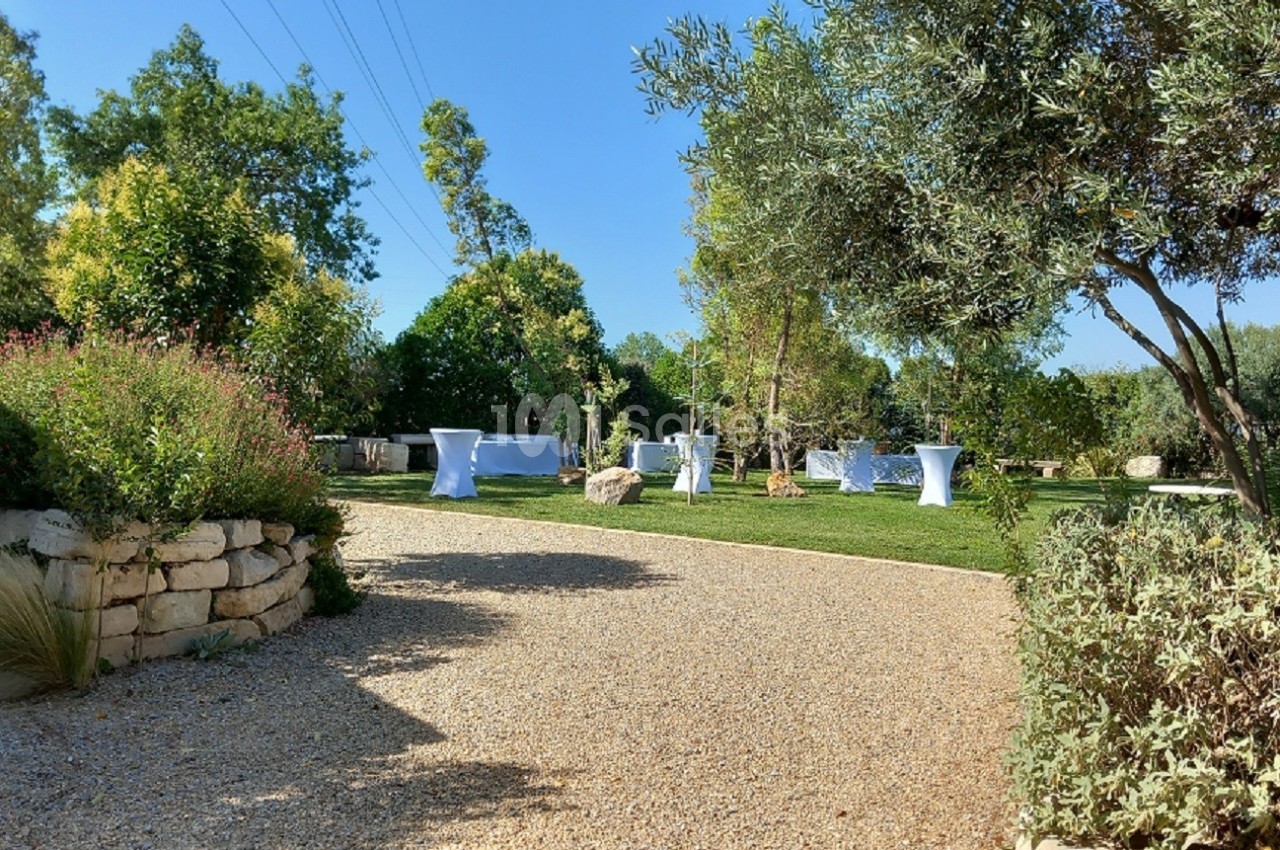 Allée en gravier menant à un jardin verdoyant avec des tables hautes blanches disposées sur une pelouse.