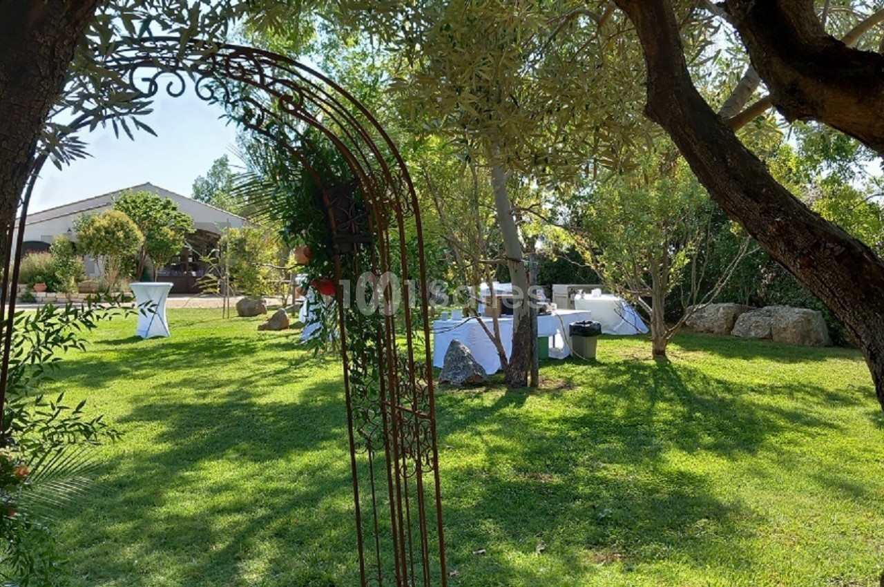 Vue d'un jardin verdoyant avec des tables dressées pour un événement, encadré par une arche métallique et des arbres.