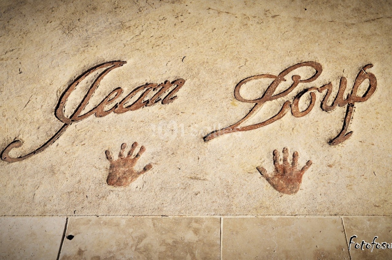 Inscription ’Jean Loup’ gravée dans une surface beige avec deux empreintes de mains en dessous.