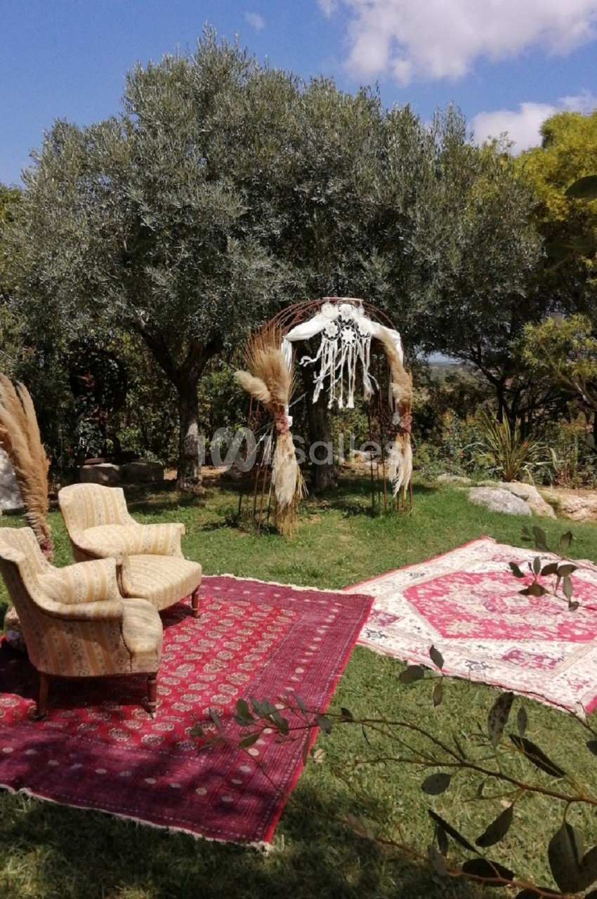 Deux fauteuils et des tapis rouges disposés sur une pelouse, sous une arche décorée de pampas et de macramé, près d'oliviers.
