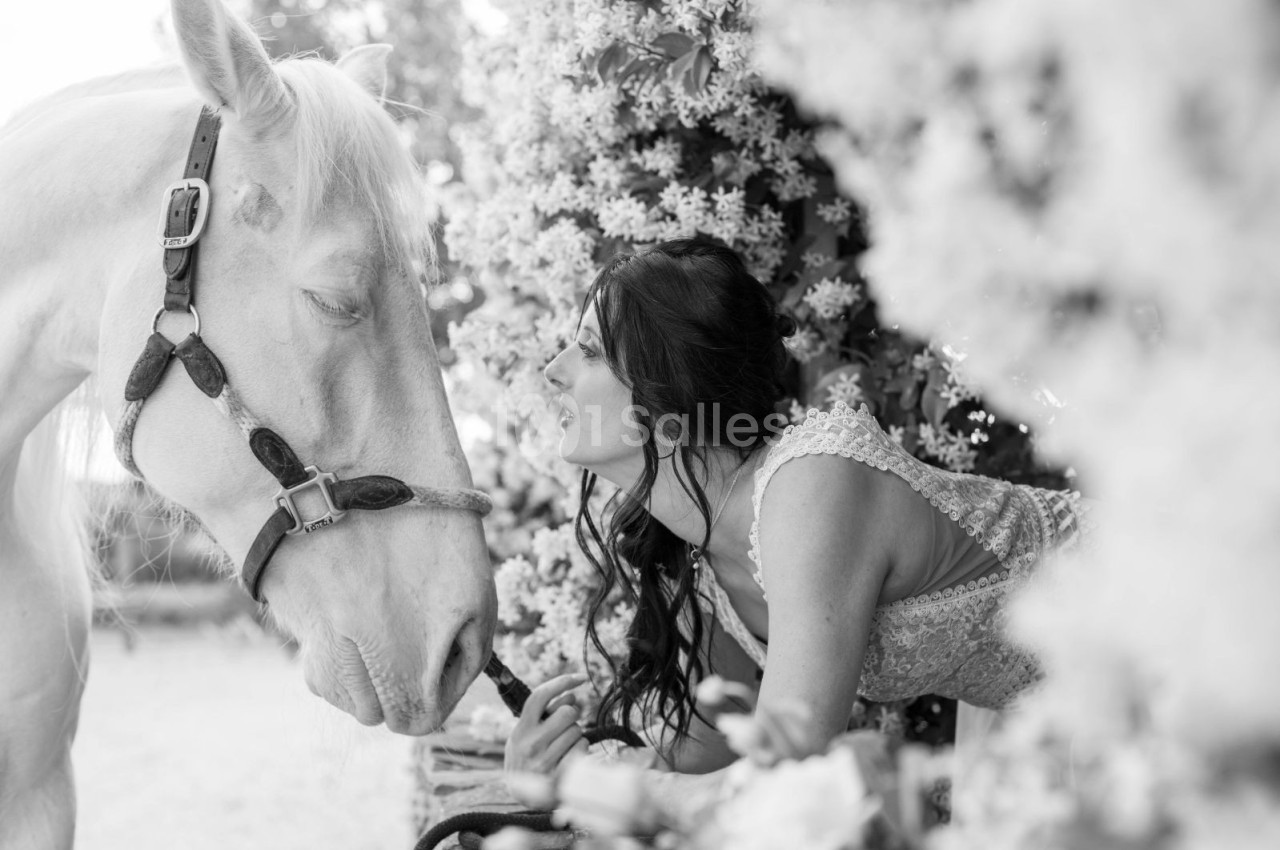 Une femme en robe légère interagit avec un cheval blanc dans un décor fleuri en noir et blanc.