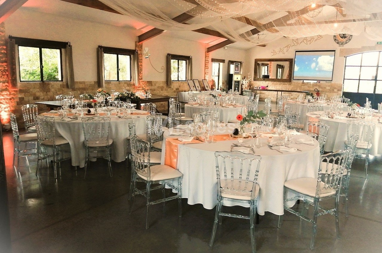Salle de réception décorée avec des tables rondes dressées, des chaises transparentes et des voilages suspendus.