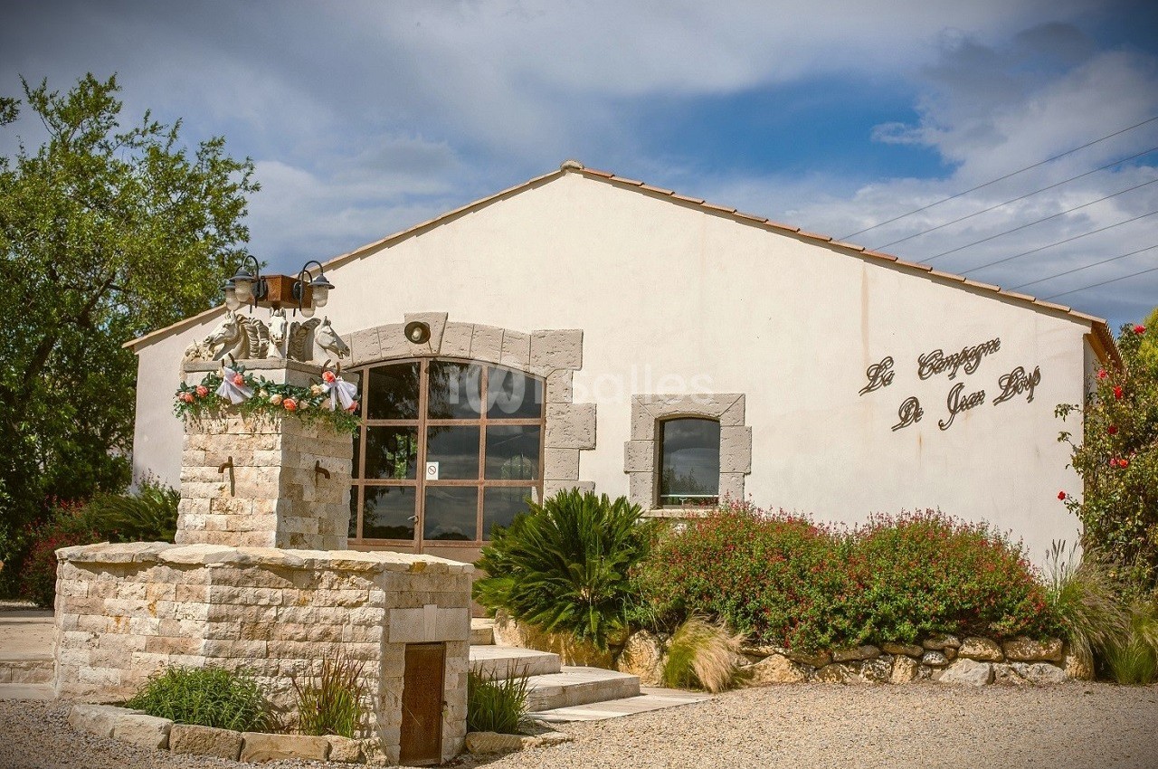 Bâtiment en pierre avec une fontaine fleurie au premier plan, entouré de végétation sous un ciel partiellement nuageux.