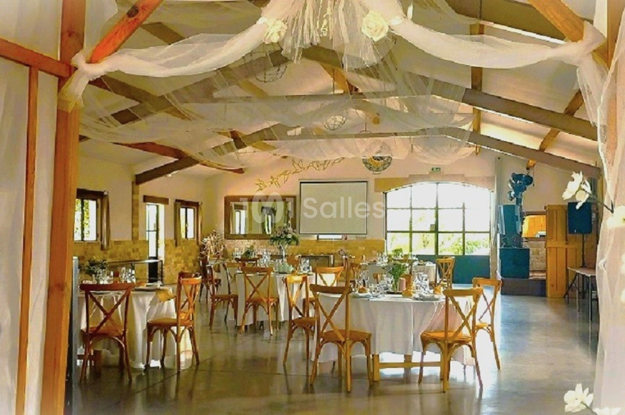 Salle de réception lumineuse décorée avec des voilages blancs, des tables rondes dressées et des fleurs blanches.