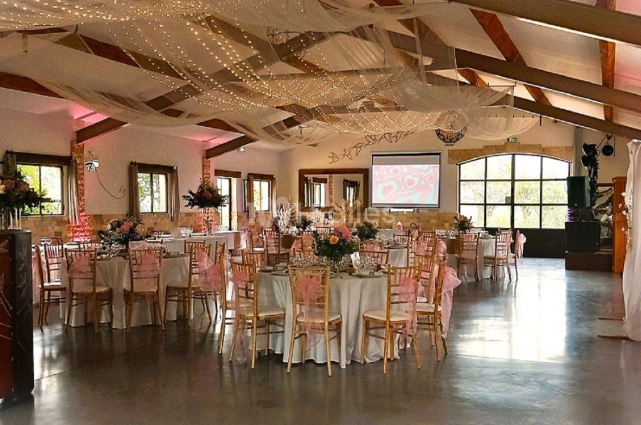 Salle de réception décorée pour un événement, avec tables rondes, chaises en bois, guirlandes lumineuses et voilages…