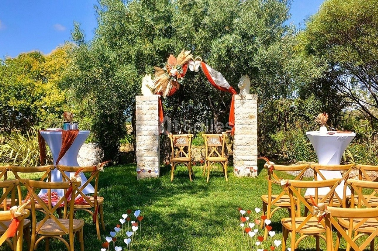 Chaises en bois disposées devant une arche décorée de fleurs et de tissus, dans un jardin ensoleillé.