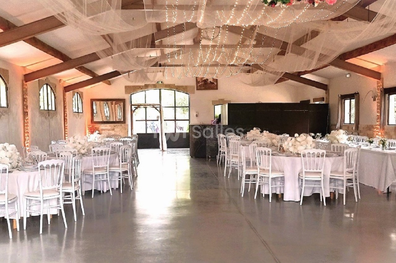 Salle de réception décorée avec des tables rondes, nappes blanches, chaises élégantes et guirlandes lumineuses suspendues.