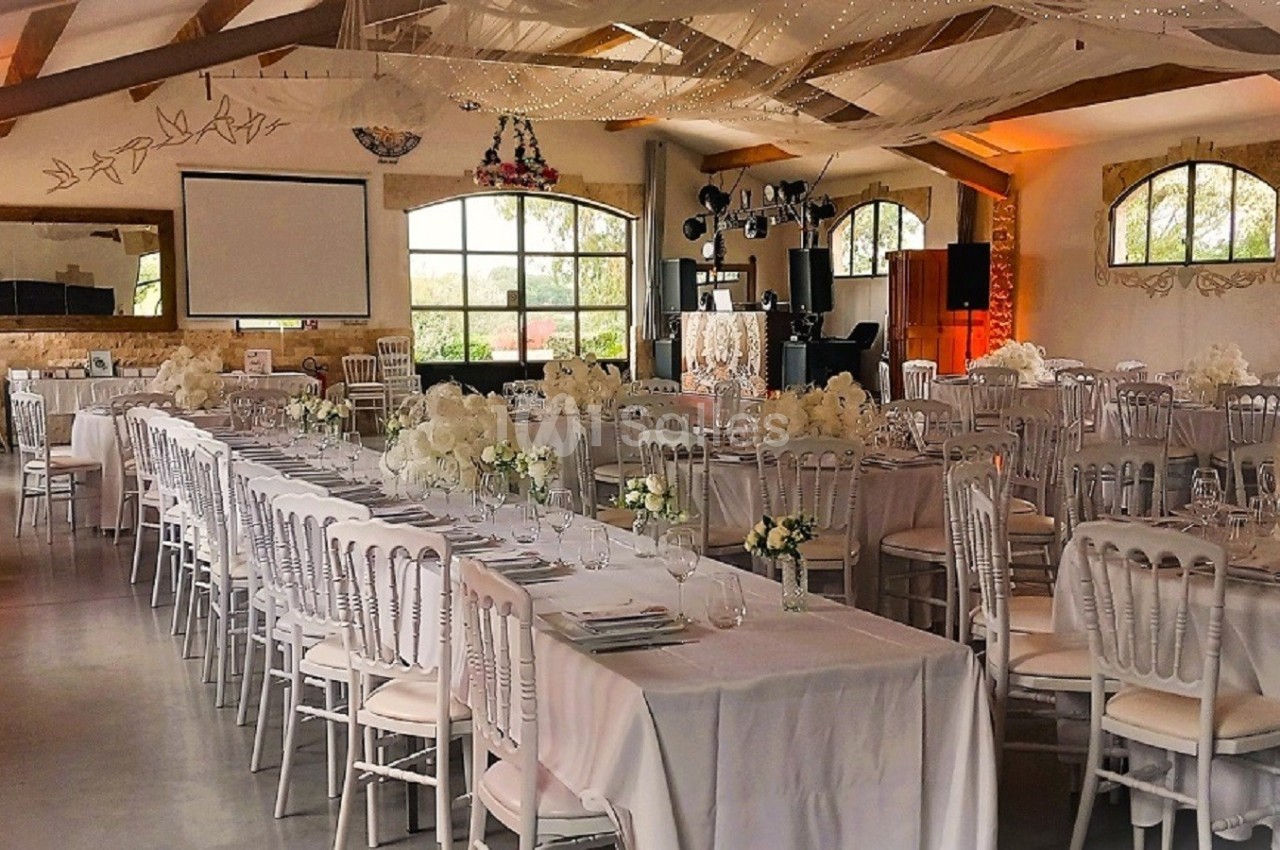 Salle de réception décorée avec des tables alignées, nappes blanches, chaises élégantes et éclairage chaleureux.