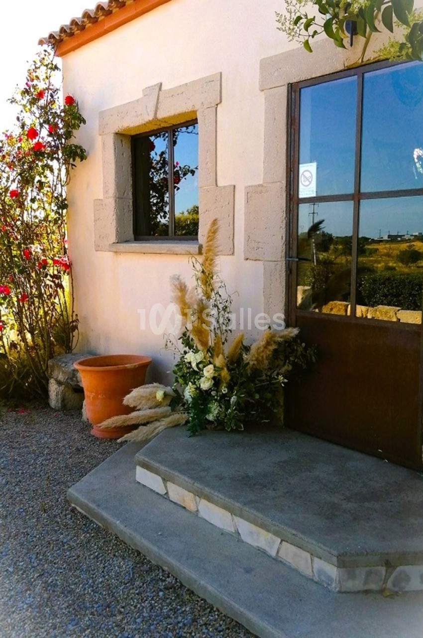 Entrée d'une maison en pierre avec porte vitrée, décorée de fleurs et de plantes, entourée de gravier et de rosiers.