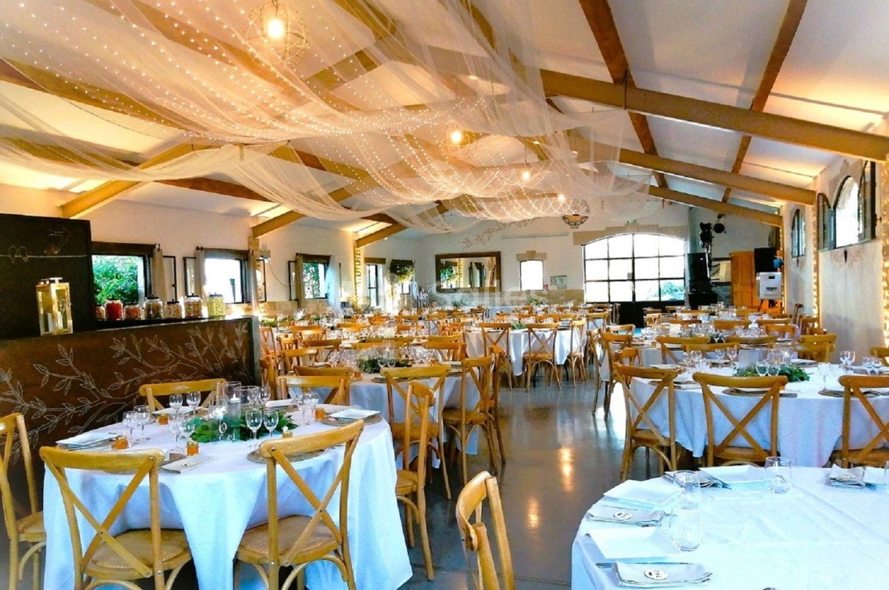 Salle de réception décorée avec des tables rondes, nappes blanches, chaises en bois et voilages suspendus au plafond.