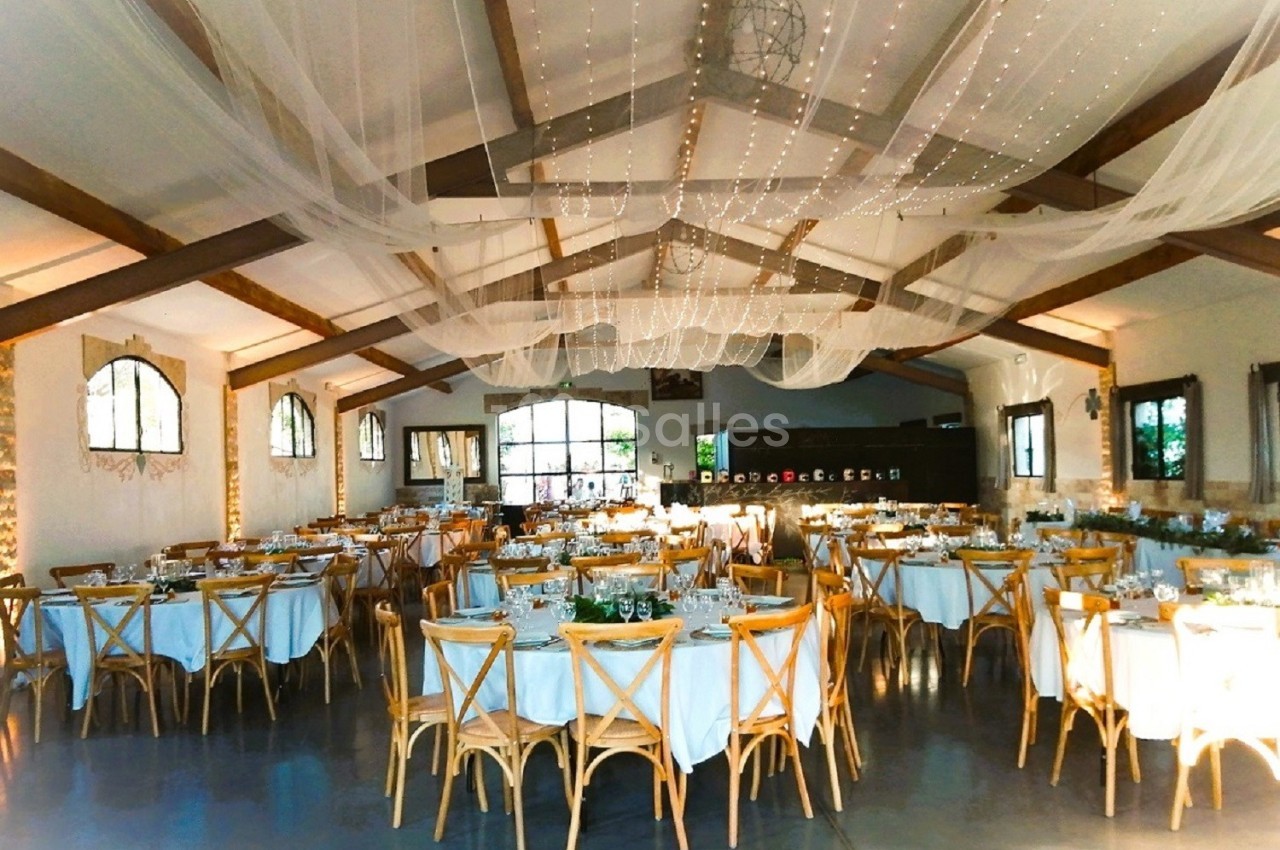 Salle de réception décorée avec des tables rondes dressées, chaises en bois et guirlandes lumineuses suspendues.