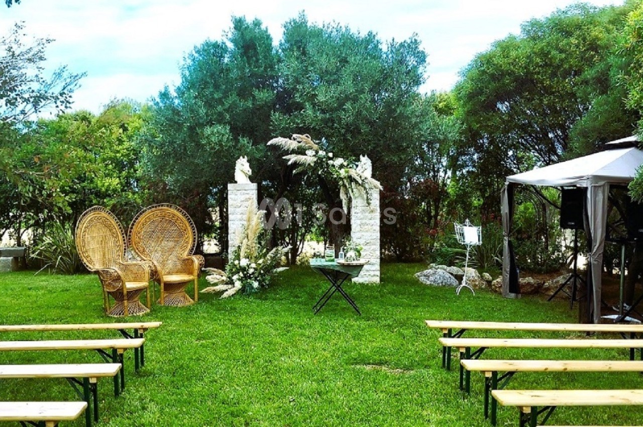 Décoration de cérémonie en plein air avec arche fleurie, fauteuils en osier et bancs en bois sur une pelouse.