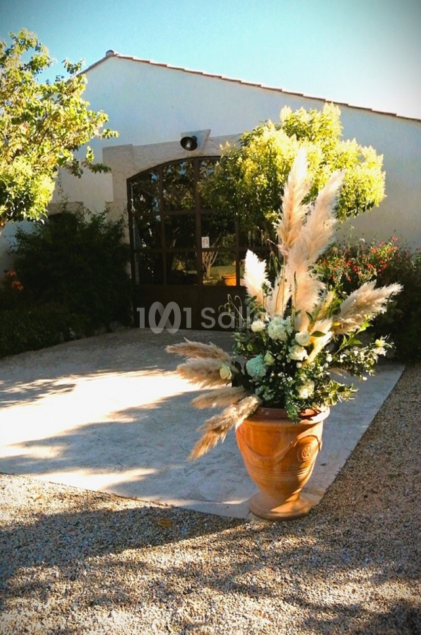Pot en terre cuite avec des fleurs et des herbes décoratives devant une bâtisse blanche entourée de végétation.